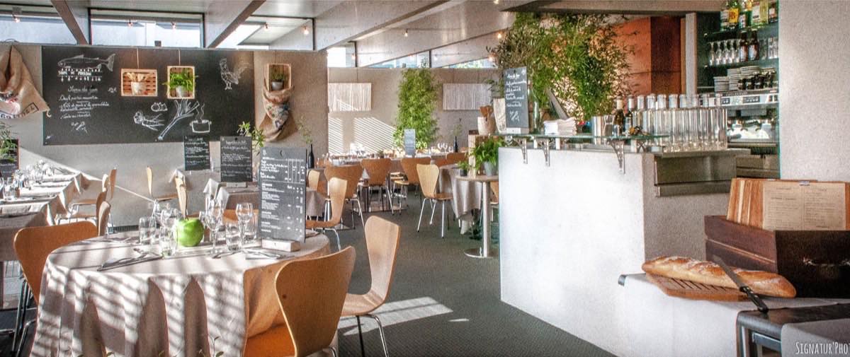 La salle du restaurant, tables rondes dressées, ardoises au mur et comptoir en arrière-plan